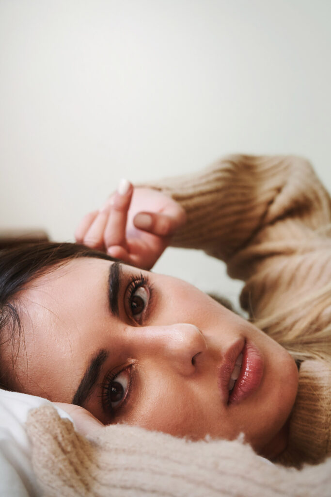 Marintia Bustos a model with striking features lies on a bed gazing intently at the camera with a soft beige sweater enveloping her.